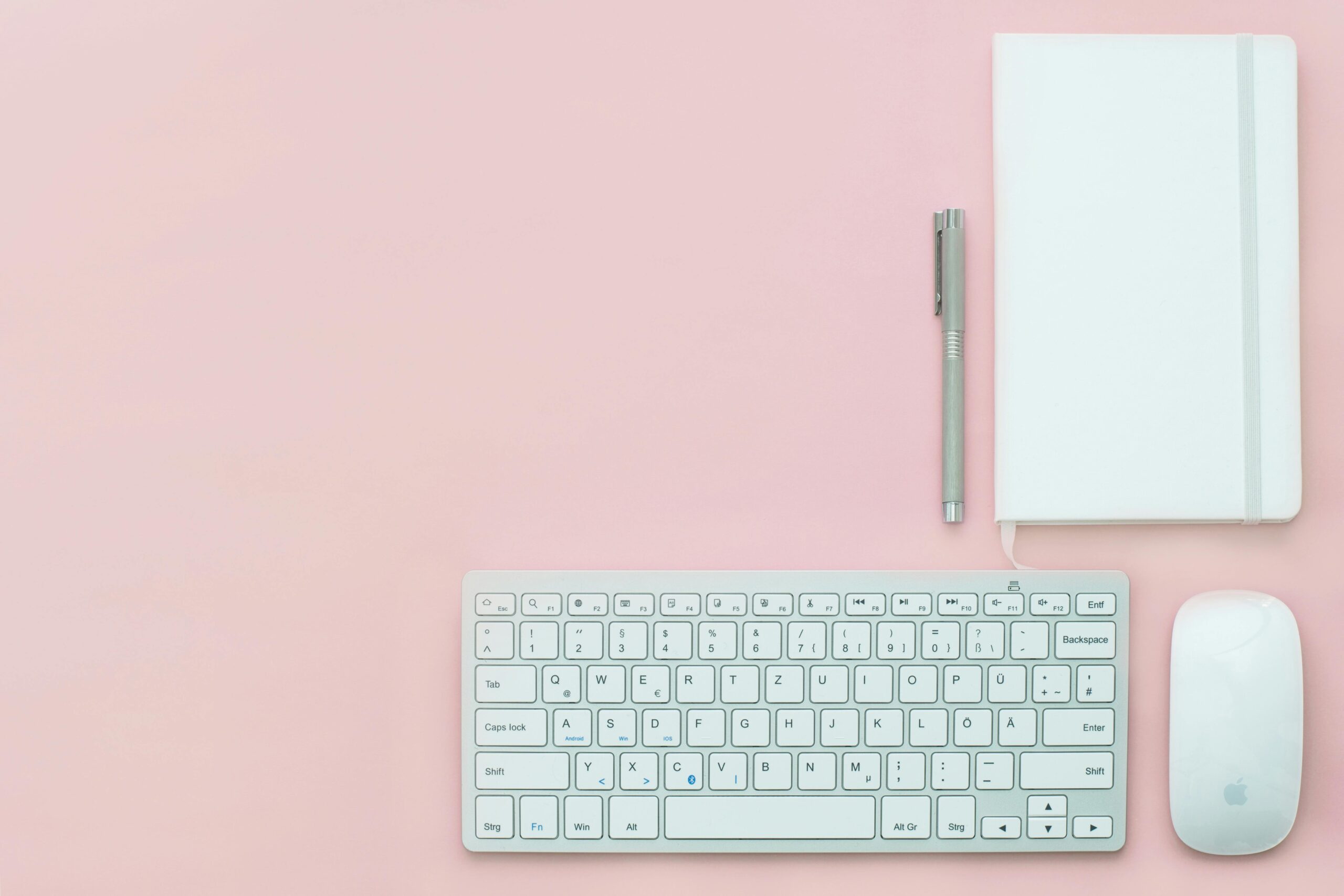 Flat lay of a minimalist workspace featuring a keyboard, notebook, and pen on a pink surface.